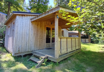 Holzhütte Chalet Bois Comfort mit überdachter Veranda im Grünen, umgeben von Bäumen und Wiese.