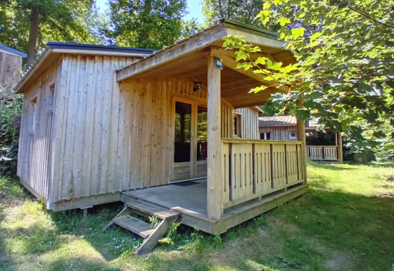 Holzhütte Chalet Bois Comfort mit überdachter Veranda im Grünen, umgeben von Bäumen und Wiese.