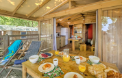 Outdoor breakfast setup on the terrace of a wooden lodge with cozy interior at Flower Camping La Chênaie, France.
