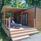 Moderne Homeflower Premium lodge med træterrasse og liggestole omgivet af grønne træer i sollys.