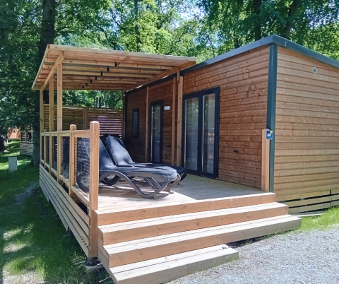 Homeflower Premium lodge met overdekt houten terras en ligstoelen, omringd door groene bomen en zon.