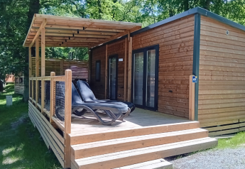 Lodge Homeflower Premium avec terrasse en bois couverte et transats, entouré d’arbres verts ensoleillés.