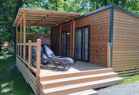 Homeflower Premium lodge met overdekt houten terras en ligstoelen, omringd door groene bomen en zon.