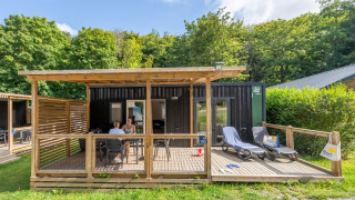 Buitenaanzicht van Homeflower Premium lodge met terras, ligzetels en mensen, omringd door natuur en bomen.