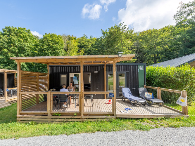 Buitenaanzicht van Homeflower Premium lodge met terras, ligzetels en mensen, omringd door natuur en bomen.
