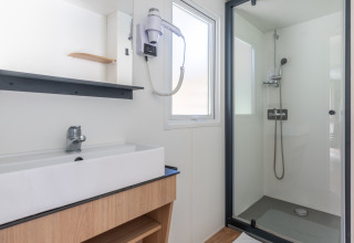 Lichte, moderne badkamer in een glampingaccommodatie met douche, wastafel, spiegel en föhn bij raam.