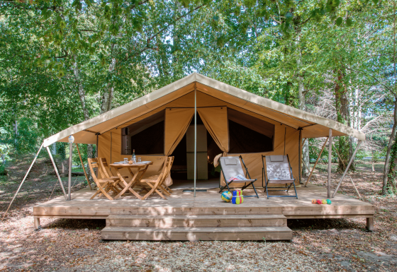 Safari tent met houten veranda, buitenzitjes en speelgoed, omringd door groene bomen in het bos.