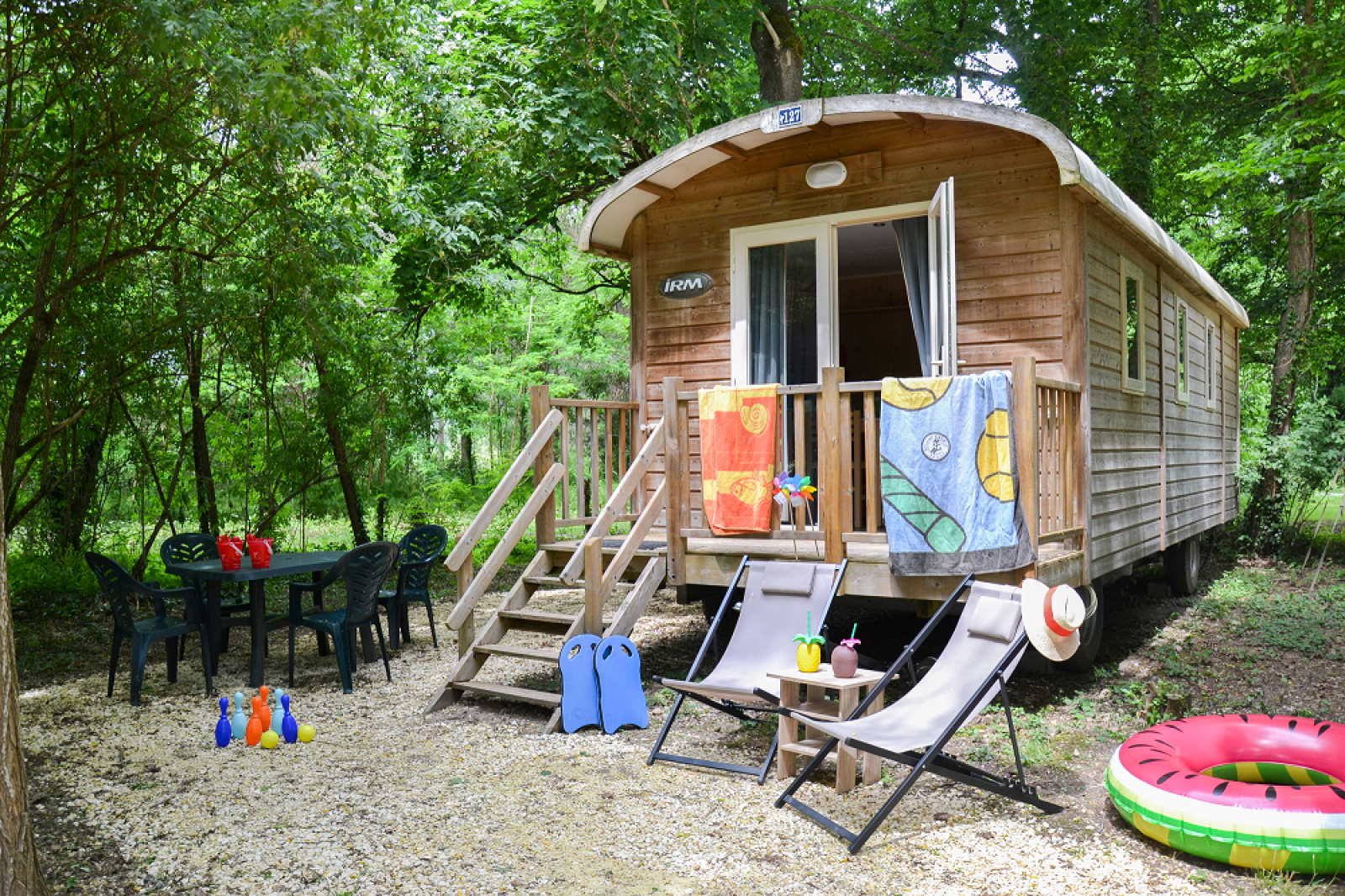 Roulotte Standard lodge in het bos met stoelen, tafel, kinderspeelgoed en kleurrijke handdoeken buiten.