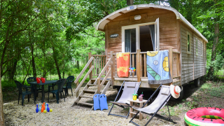 Roulotte Standard lodge in het bos met stoelen, tafel, kinderspeelgoed en kleurrijke handdoeken buiten.