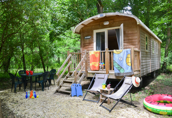 Roulotte Standard lodge in het bos met stoelen, tafel, kinderspeelgoed en kleurrijke handdoeken buiten.