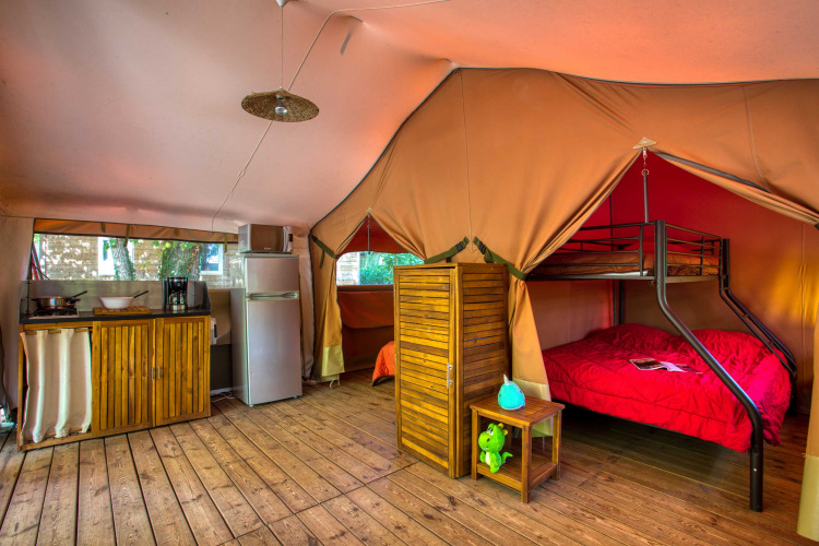 Indretning af et hyggeligt safari-telt med køkken, køleskab, trægulv og køjeseng på Flower Camping La Sagne.