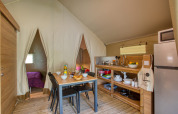 Vista interior de una tienda safari con cocina, mesa de comedor y dormitorio en Flower Camping La Sagne, Francia.