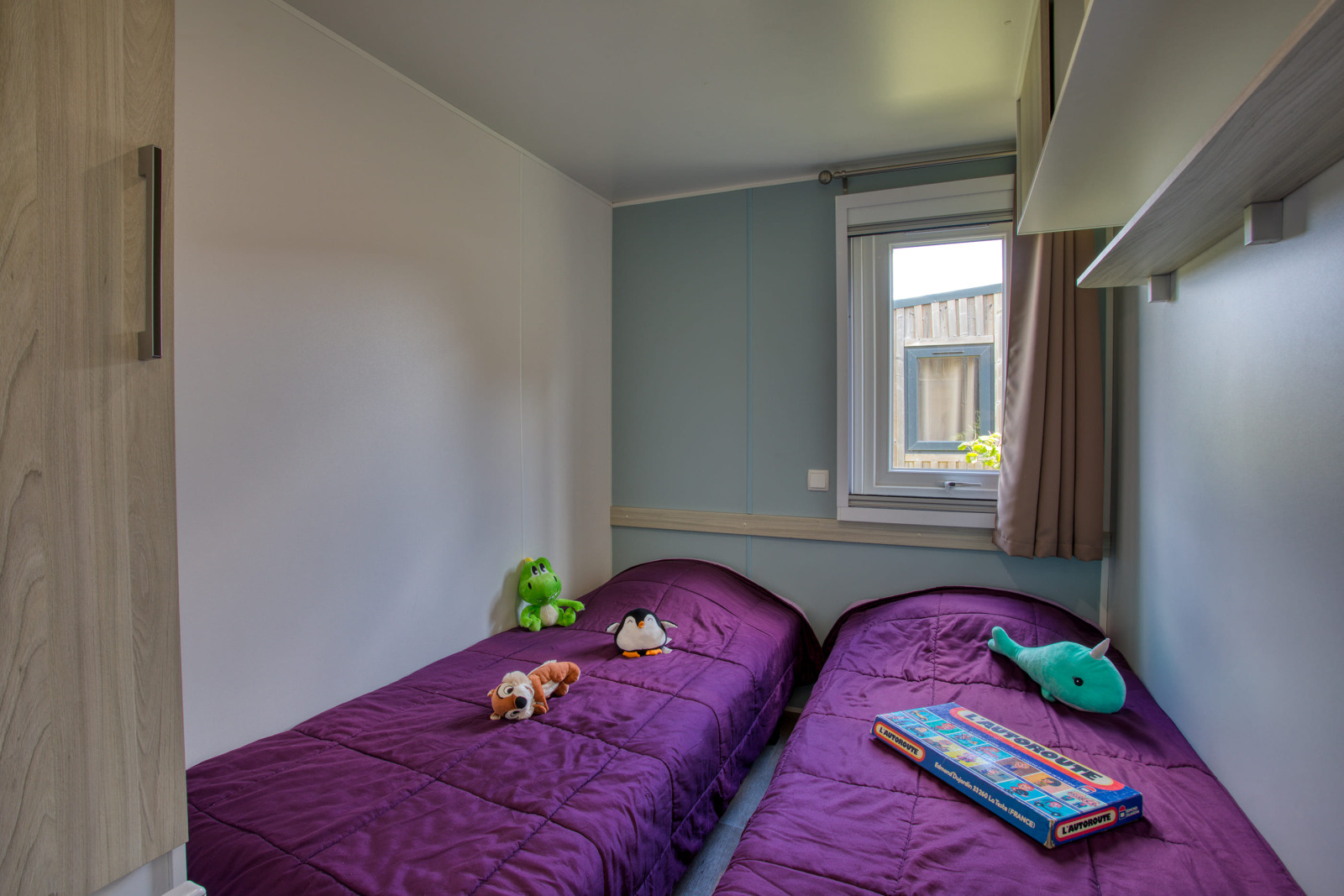 Twee eenpersoonsbedden met paarse dekbedden, knuffels en een raam in Stacaravan Campbell Premium, Frankrijk.