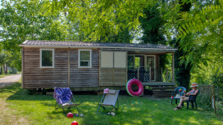 A mobile home called Stacaravan Cayuga Confort with a wood exterior, outdoor seating, toys, and lush greenery.