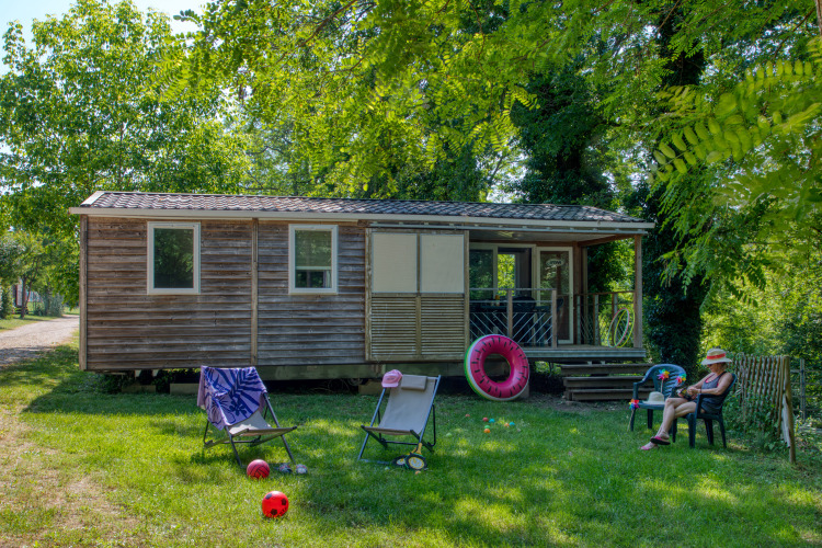A mobile home called Stacaravan Cayuga Confort with a wood exterior, outdoor seating, toys, and lush greenery.