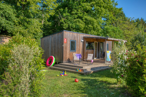 Wooden lodge Stacaravan Arlequin Premium surrounded by lush trees, toys on the grass and a sunny porch.