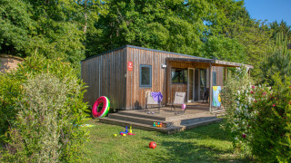 Holzlodge Stacaravan Arlequin Premium, umgeben von Bäumen, Holzterrasse und Spielzeug im grünen Garten.