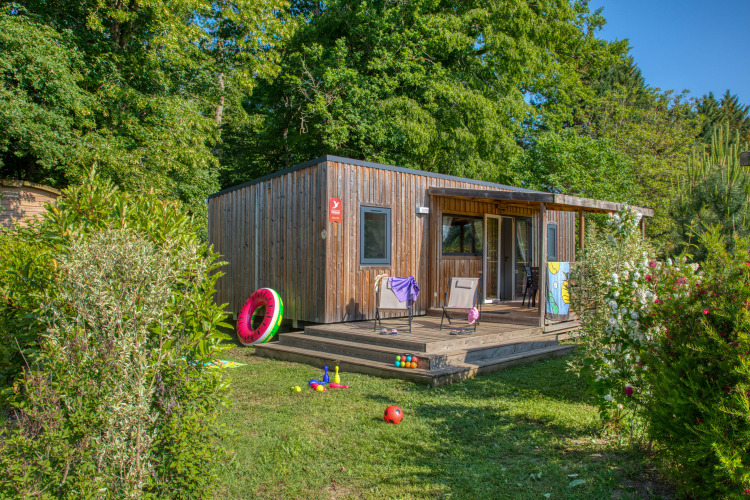 Chalet en bois Stacaravan Arlequin Premium entouré d’arbres verts, jouets colorés et terrasse ensoleillée.