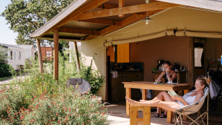 Two people relax at a wooden table and chair under the canopy at a Ecolodge Cotton Toilée CONFORT safari tent.