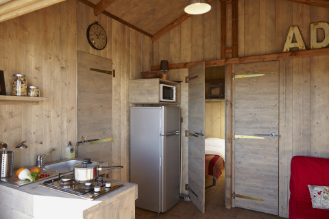 Interior view of a wooden cabin with kitchenette, stove, sink, fridge, and open door to bedroom.