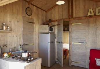 Binnenzicht van een houten hut met keukenhoek, kookplaat, gootsteen, koelkast en open deur naar slaapkamer.