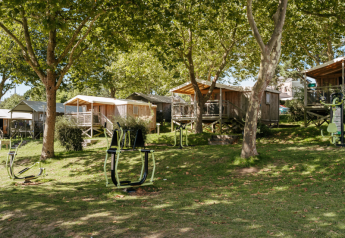Gemütliche Holzhütten im Grünen mit Outdoor-Fitnessgeräten, von Bäumen beschattet im Sonnenlicht.