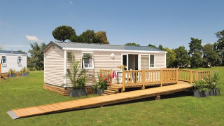 Modern glampingverblijf met houten terras en oprijlaan op een grasveld onder een blauwe hemel.