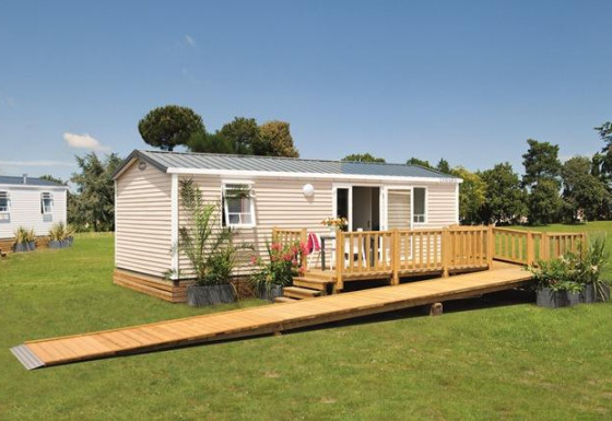 Modern glamping-verblijf met houten terras en oprit op een grasveld onder een heldere blauwe lucht.