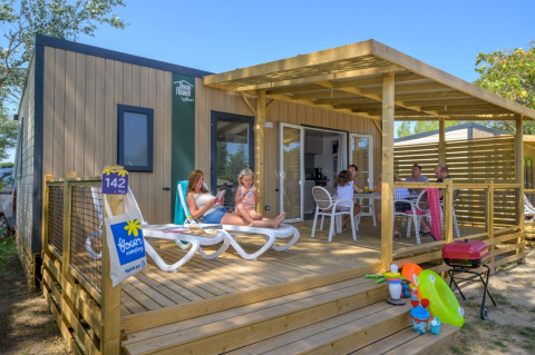 Family relaxing on the wooden terrace of Homeflower Premium lodge with toys and a barbecue in the foreground.