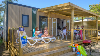 Family relaxing on the wooden terrace of Homeflower Premium lodge with toys and a barbecue in the foreground.