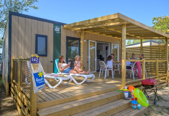 Family relaxing on the wooden terrace of Homeflower Premium lodge with toys and a barbecue in the foreground.