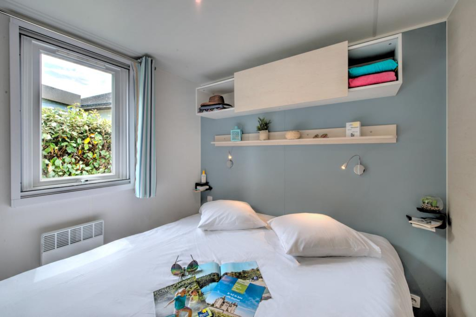 Lichte glamping slaapkamer met tweepersoonsbed, groot raam, wandplanken en moderne inrichting.