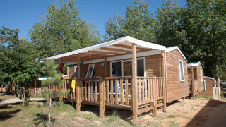Cabina Stacaravan Confort con veranda in legno e tenda bianca, circondata da alberi in una giornata di sole