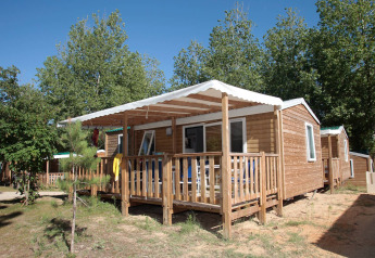 Cabaña Stacaravan Confort con porche de madera y toldo blanco, rodeada de árboles en un día soleado