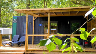 Photo of a glamping cabin with wooden deck, lounge chairs, and lush greenery in a forest setting.