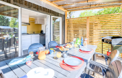 Coin repas extérieur au glamping avec table dressée, boissons, viennoiseries sur terrasse ensoleillée avec barbecue.