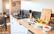 Moderne glampingkeuken met gasfornuis, spoelbak, saladekom en een eettafel in een lichte ruimte.