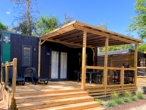 Hébergement glamping moderne avec façade noire, grande terrasse en bois et coin salon ombragé.