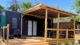 Hébergement glamping moderne avec façade noire, grande terrasse en bois et coin salon ombragé.