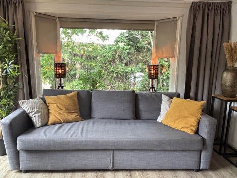 Grijze sofa met gele kussens bij grote ramen in Cozy Lodge, Bungalowpark Het Verscholen Dorp, Nederland.