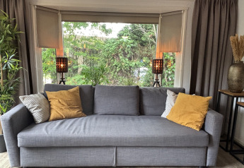 Divano grigio con cuscini gialli davanti a grandi finestre al Cozy Lodge, Bungalowpark Het Verscholen Dorp, Paesi Bassi.