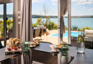Frühstückstisch mit Brötchen, Marmelade und Gurkenwasser in einem Ferienpark mit Blick auf Pool und Meer.