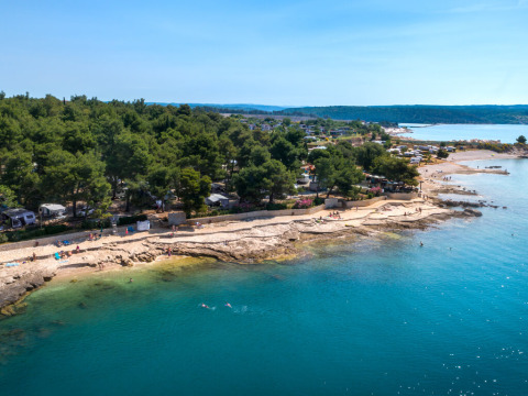 Kustcamping en rotsachtige strand bij Novigrad, Istrië, Kroatië, met helderblauw water en veel bomen.