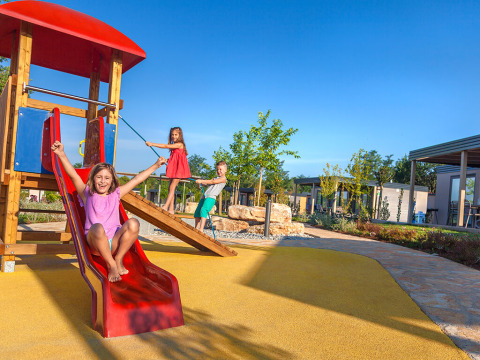 Drei Kinder spielen an einer Rutsche auf einem Spielplatz vor den Maro Home Chalets bei sonnigem Wetter.