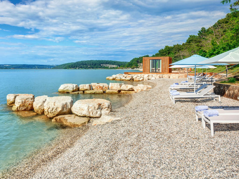 Kiezelstrand met ligbedden, parasols en een luxe safaritentgebouw aan de rustige zee.