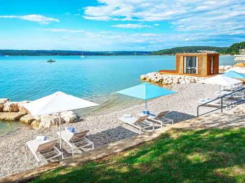 Transats et parasols sur une plage de galets ensoleillée près d'une cabane moderne au bord de l'eau.