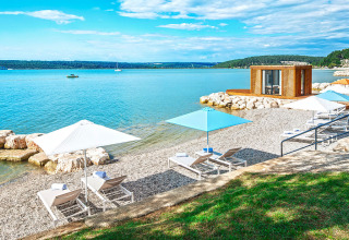 Transats et parasols sur une plage de galets ensoleillée près d'une cabane moderne au bord de l'eau.