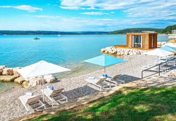 Lettini e ombrelloni su una spiaggia di ciottoli, vicino a una moderna cabina e acque calme.