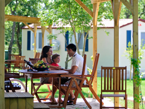 Famiglia seduta a un tavolo di legno davanti a lodge glamping immersi nella natura verdeggiante.