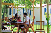 Familie sitzt an Holztisch unter Pavillon vor modernen Glamping-Unterkünften im Grünen.